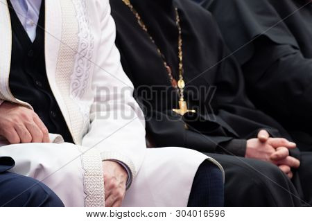 A Muslim Mullah In White Clothes And A Christian Priest In A Black Cassock Are Sitting Next To Each 