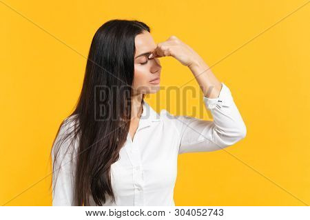 Portrait Of Exhausted Young Woman In White Casual Shirt Keeping Eyes Closed, Putting Hand On Nose Is
