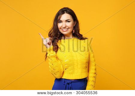 Portrait Of Smiling Young Woman In Sweater Blue Trousers Looking Camera Pointing Index Finger Aside 