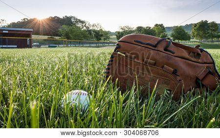 Baseball And Glove On Grass In Morning Dew Near Dugout In Early Morning With Sunburst