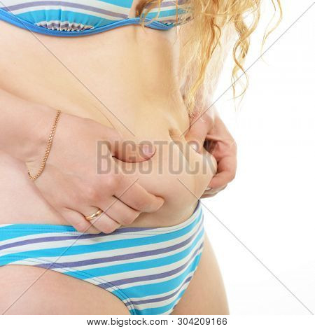 Overweight young woman shows belly fat, isolated over white background. Overeating xl size girl dressed in swimsuit unhappy with her figure and overweight