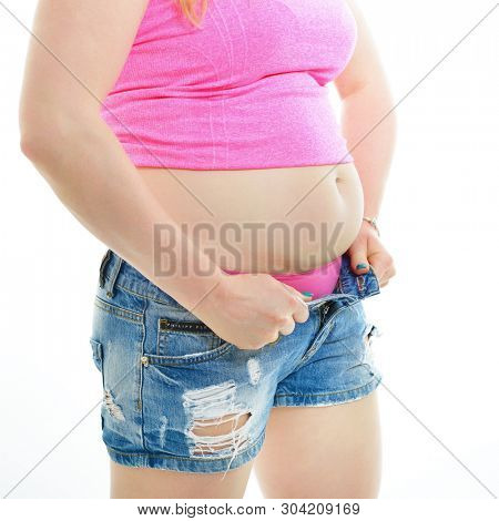 Overweight young woman, isolated over white background. Overeating xxl size girl dressed in denim blue shorts and a pink t-shirt trying to zip up shorts