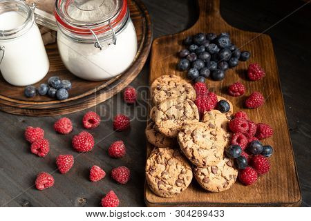 Two Wooden Platters With Chcocolate Biscuits, Raspberries, Blueberries, Milk And Flour.