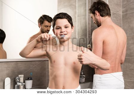 Son Brushing Teeth Near Father And Showing Thumb Up In Bathroom