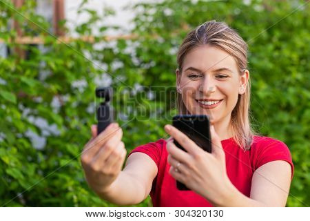 Young Blonde Woman Holding Phone And Osmo Camera For Vlogging Outdoor , Green Background