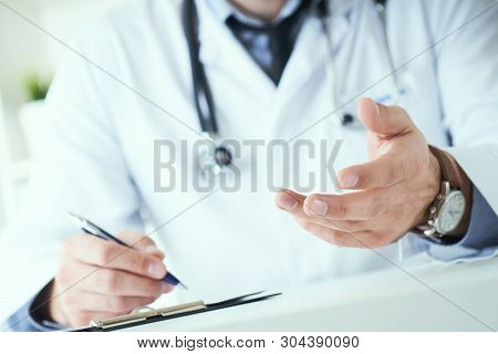 Male Doctor Making Welcome Gesture, Politely Inviting Patient To Sit Down In Medical Office. Photo W