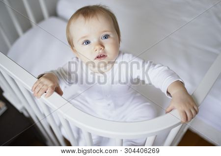 Adorable Baby Girl In Co-sleeper Crib Attached To Parents Bed. Little Kid Sitting In Cot