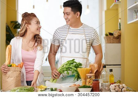 Young Asian Couple In Love Standing At Kitchen Table With Various Fresh Pruducts They Bought In Groc