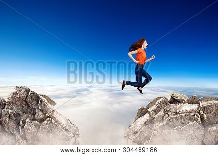 Redhead Woman Jumps Through The Gap Between Hills Over Cliff On Blue Sky Background.