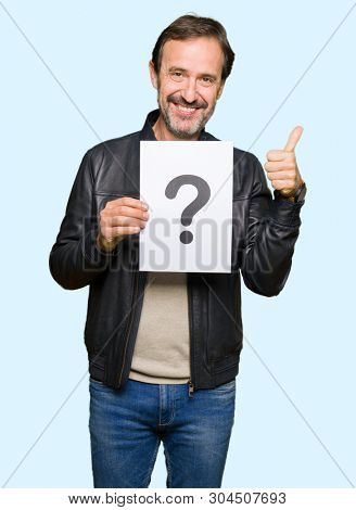 Handsome middle age man holding paper with question mark happy with big smile doing ok sign, thumb up with fingers, excellent sign