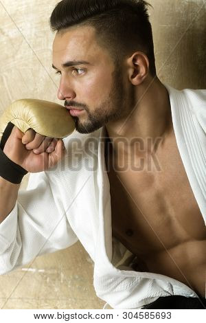 Attractive Guy Siting On Sofa And Thinking About Fight. Man In Kimono And Boxing Gloves Is Preparing
