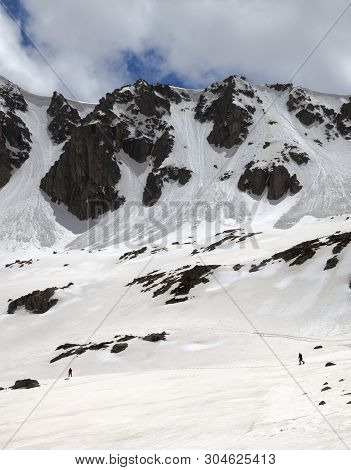 High Mountains With Snow Cornice And Avalanche Trail, Snowy Plateau And Two Small Silhouette Of Hike