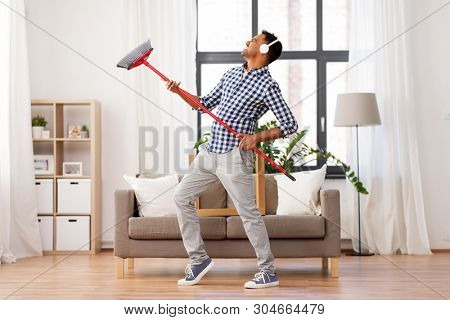 cleaning, housework and housekeeping concept - indian man in headphones with broom sweeping and having fun at home