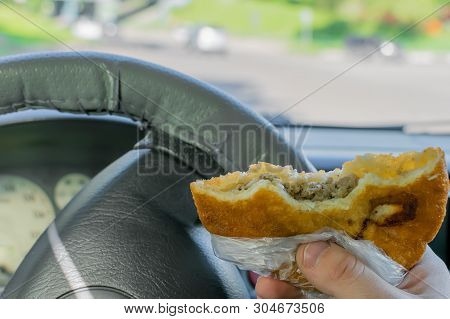 Pie, Belyash With Meat In The Hand Of The Driver In The Car On The Background Of The Steering Wheel 