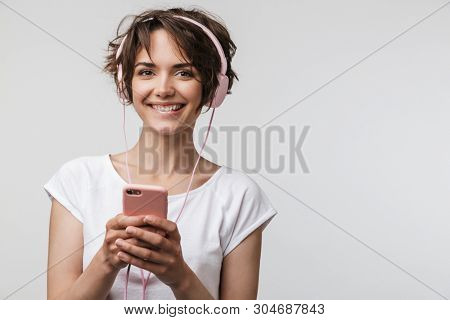 Image of optimistic woman in basic t-shirt holding smartphone while listening to music with headphones isolated over white background