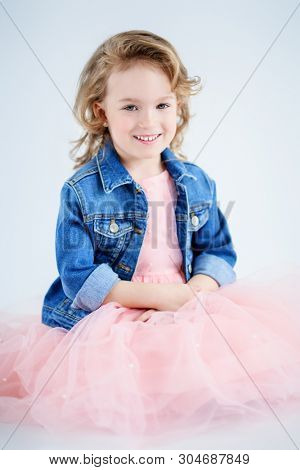 A portrait of a pretty girl in a pink dress and a denim jacket posing in the studio over the white background. Kids, fashion, beauty.