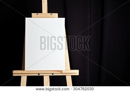 White Blank Cotton Canvas Stands On A Wooden Artistic Easel On Black Curtain Background. Vertical Re