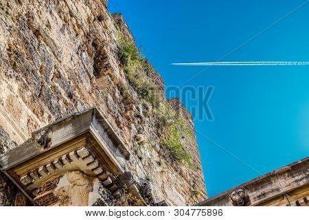 The Plane Leaving A Condensation Trail Flies High Above The Gates Of Hadrian. Adrian Gate, Antalya L