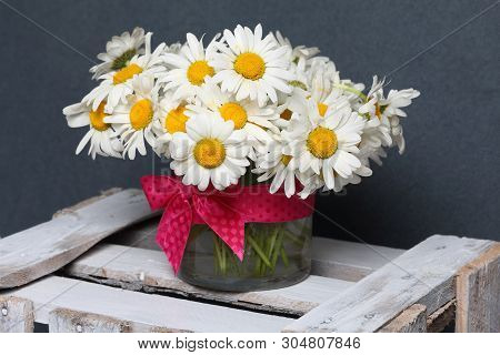 A Bouquet Of Field Daisies In A Glass Vase. On The  Wooden Box. Genus Of Perennial Flowering Plants