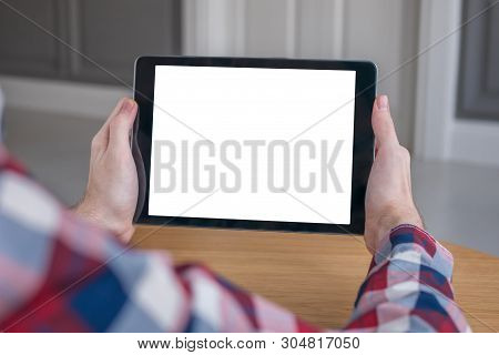 Man Sitting At Wooden Table And Looking At Black Digital Tablet Computer Device With White Blank Scr