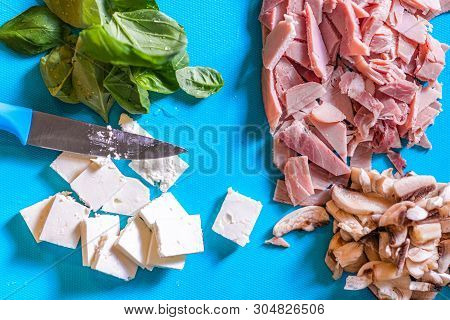 Cut Up Ingredients Such As Ham, Mushroom, Cheese And Basil On A Blue Plastic Chopping Board With A S