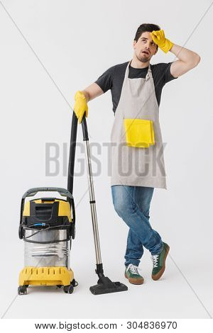 Full length of a handsome brunette houseman wearing apron standing isolated over white background, cleaning with vacuum cleaner