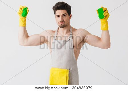 Handsome brunette houseman wearing apron standing isolated over white background, holding sponges