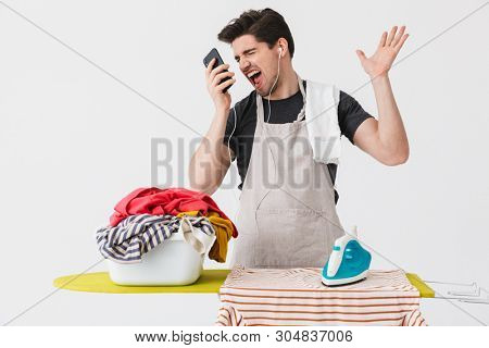 Handsome angry brunette houseman wearing apron standing isolated over white background, ironing clothes on a board, talking on mobile phone
