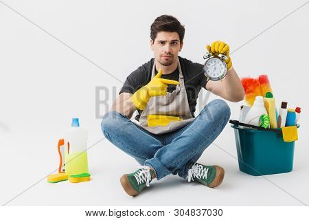 Handsome brunette houseman wearing apron sitting with bucket full of detergents isolated over white background, holding alarm clock