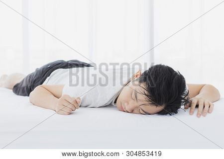 Asian Man Lie Prone On Bed, He In Pajamas In Bedroom.