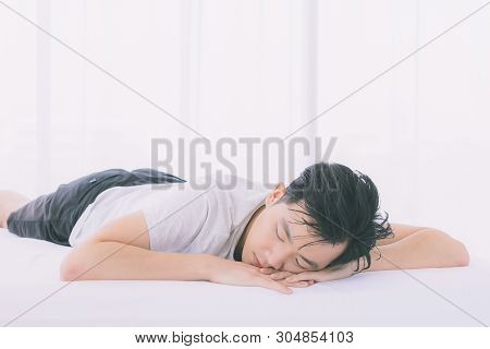 Asian Man Lie Prone On Bed, He In Pajamas In Bedroom.