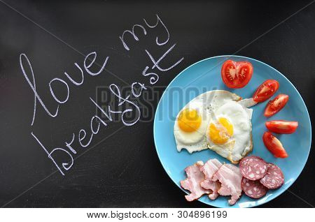 Two Fried Eggs With Fresh Tomatoes, Bacon And Sausage On The Round Blue Clay Plate On The Black Chal