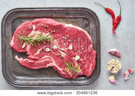 Raw Steak. Meat With Garlic, Pepper And Rosemary Im Metal Tray. Top View On Light Stone Background.