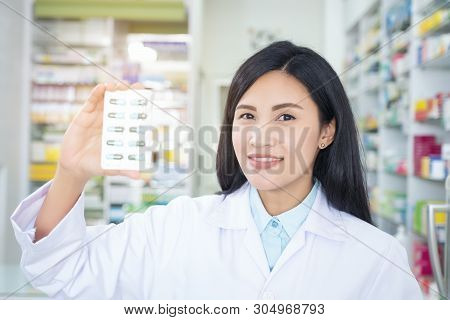 Pharmacist Holds A Packet Of Pills In Her Hands. Then She Shows A Thumbs Up. A Woman Is Standing In 