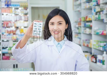 Pharmacist Holds A Packet Of Pills In Her Hands. Then She Shows A Thumbs Up. A Woman Is Standing In 