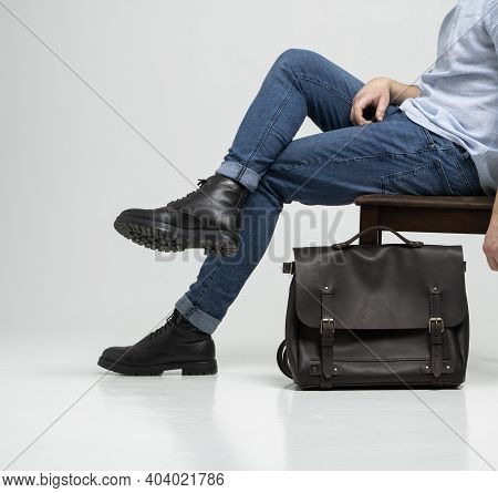 Man In A Blue Jeans And Black Boots Sits On A Chair With A Brown Mens Shoulder Leather Bag For A Doc