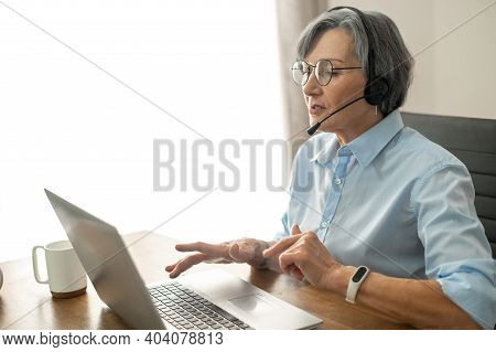 Grey-headed Mature Senior Female Office Worker Sitting At The Desk With A Laptop, Wearing Glasses, H