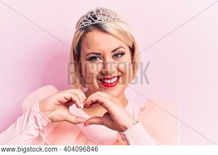 Beautiful blonde plus size woman wearing pincess tiara with diamonds over pink background smiling in love doing heart symbol shape with hands. Romantic concept.