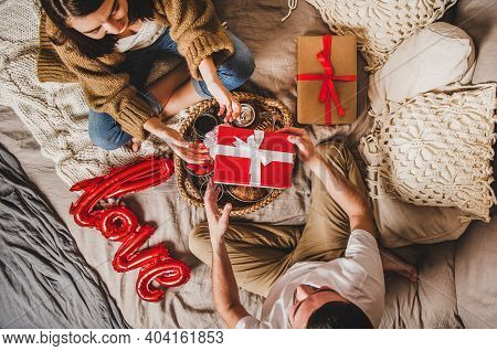 Young Happy Couple Sitting On Blankets In Bed At Home And Giving Presents In Boxes Over Breakfast An
