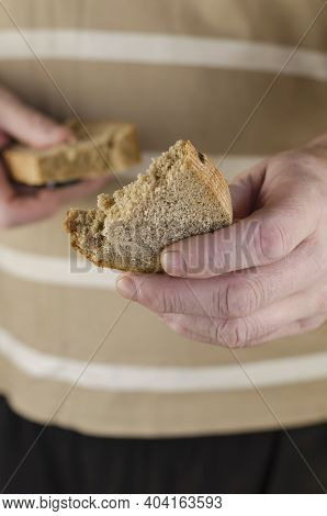A Man's Hand Holds Out Half A Slice Of Rye Bread To The Camera. In The Background, The Hand Holds Th