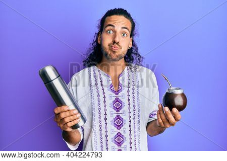 Young handsome man with long hair drinking mate infusion puffing cheeks with funny face. mouth inflated with air, catching air. 