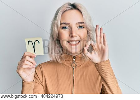 Young blonde girl holding paper with aries zodiac sign doing ok sign with fingers, smiling friendly gesturing excellent symbol 