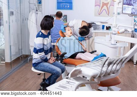 Little Girl In The Course Of Treatment For Caries Sitting On Dental Chair Wearing Bib. Dentistry Spe