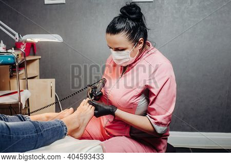 The Master Makes A Pedicure In The Beauty Salon. The Girl In A Blue Mask And Gloves Makes Cosmetic P