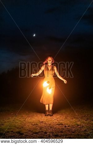 Graceful elegant red haired female fireshow performer making fire dance with two metal fans lit with flames on night mountains meadow
