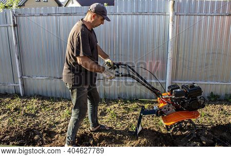 Male In The Spring Plows The Earth In Working Clothes With A Motoblock, Walk-behind Tractor