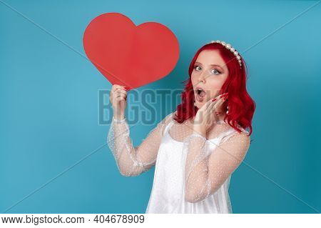 Shocked Dumbfounded Young Woman With Open Mouth In White Dress And Red Hair Holding A Big Red Paper 