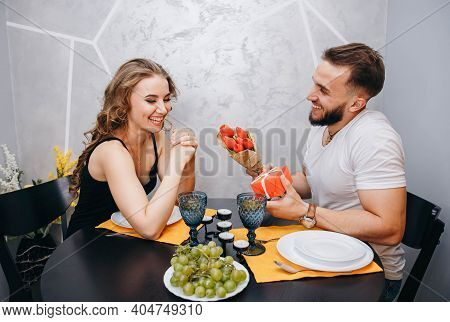 Beautiful Couple Sitting At Table With Flower Bouquet And Looking At Each Other During Romantic Date