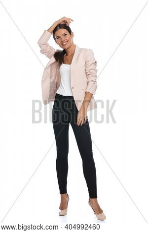attractive young woman wearing pink jacket, holding hand over head and posing in a fashion manner, standing isolated on white background in studio, full body