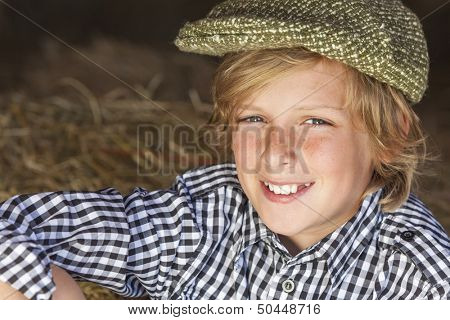 Young happy smiling blond boy child aged about twelve or early teenager wearing a plaid shirt and flat cap sitting on hay or straw bales